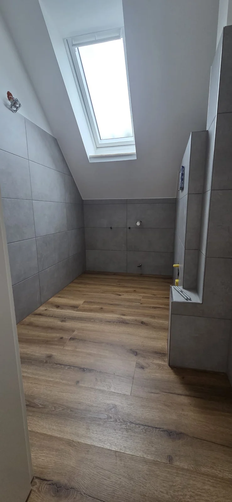 Sanitärraum unter der Dachschräge mit grauen Wandfliesen und Boden in Holzoptik